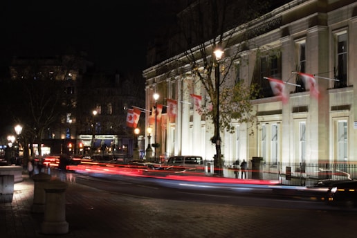 A nighttime city scene featuring a well-lit building adorned with Canadian flags. The street is busy with motion blur from passing vehicles, creating streaks of light. Trees and lampposts line the road, and a sense of urban life is evident.
