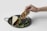 A hand holding a taco filled with meat, positioned above a dark plate with another taco on it. Fresh green cilantro is scattered around, adding a touch of color.
