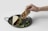A hand holding a taco filled with meat, positioned above a dark plate with another taco on it. Fresh green cilantro is scattered around, adding a touch of color.