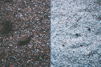 The image displays a split view of two different textures of gravel or pebbles. The left side features darker, multicolored stones with a few larger rocks, creating a rich, earthy texture. The right side contrasts with lighter-colored, predominantly white pebbles, offering a more uniform and subtle appearance.