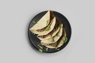 A black plate holds a stack of four tacos filled with beef and garnished with fresh cilantro leaves, set against a light gray background.