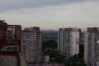 A view of a series of high-rise residential buildings with visible balconies, surrounded by greenery at the lower levels. The city skyline is faintly visible in the distance under a cloudy sky. Various power lines crisscross the image, adding to the urban setting.