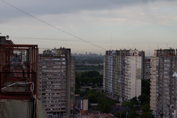 A view of a series of high-rise residential buildings with visible balconies, surrounded by greenery at the lower levels. The city skyline is faintly visible in the distance under a cloudy sky. Various power lines crisscross the image, adding to the urban setting.