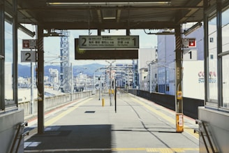 An empty urban train platform with overhead signage showing directions and train numbers. The platform has yellow safety lines marking the edges, and the cityscape with buildings and distant mountains is visible in the background. There are poles with emergency signaling and information signs on both sides.