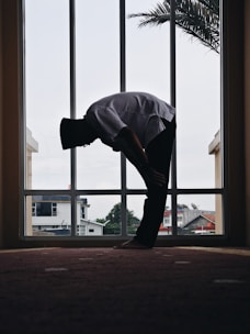 A person is silhouetted against a large window, bending forward in a prayer position. Outside the window, there are houses and trees visible under an overcast sky. The room has a minimalist design with a carpeted floor, and a palm frond is partially visible through the window.