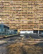 A large residential building with numerous yellow balconies is seen, each having air conditioning units installed. In the foreground, there's a wet, reflective ground surface of a parking area with some visible puddles. The parking lot has a fence along the left side and some yellow lines marking the spaces.