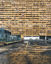 A large residential building with numerous yellow balconies is seen, each having air conditioning units installed. In the foreground, there's a wet, reflective ground surface of a parking area with some visible puddles. The parking lot has a fence along the left side and some yellow lines marking the spaces.
