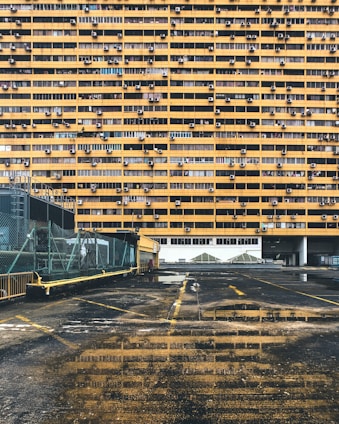 A large residential building with numerous yellow balconies is seen, each having air conditioning units installed. In the foreground, there's a wet, reflective ground surface of a parking area with some visible puddles. The parking lot has a fence along the left side and some yellow lines marking the spaces.