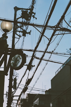 A network of black utility cables stretches across the sky with a streetlight featuring a round bulb and a power pole supporting various electrical equipment. The background includes the outlines of buildings and a clear sky.
