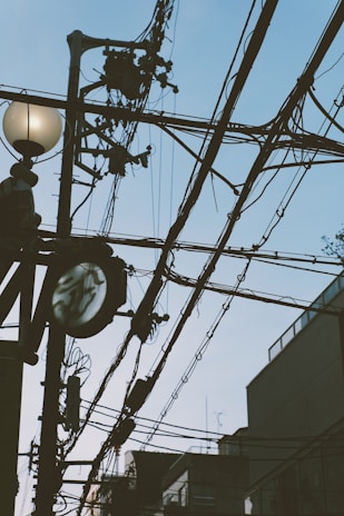 A network of black utility cables stretches across the sky with a streetlight featuring a round bulb and a power pole supporting various electrical equipment. The background includes the outlines of buildings and a clear sky.