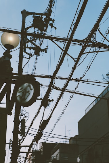 A network of black utility cables stretches across the sky with a streetlight featuring a round bulb and a power pole supporting various electrical equipment. The background includes the outlines of buildings and a clear sky.