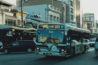 A bus drives through an urban area with multiple vehicles around it. There are buildings with varying architectural styles lining the street, along with visible traffic lights and overhead wires.