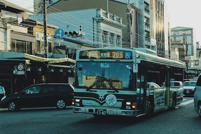 A bus drives through an urban area with multiple vehicles around it. There are buildings with varying architectural styles lining the street, along with visible traffic lights and overhead wires.