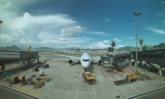 A large airplane is stationed at an airport gate, with jetways connected to it. The tarmac is busy with ground handling vehicles and stacked cargo pallets. There are distant mountains and a control tower visible in the background under a partly cloudy sky.