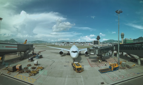 A large airplane is stationed at an airport gate, with jetways connected to it. The tarmac is busy with ground handling vehicles and stacked cargo pallets. There are distant mountains and a control tower visible in the background under a partly cloudy sky.