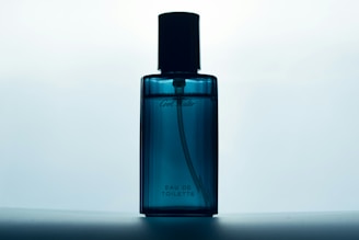 A blue glass bottle of Eau de Toilette stands against a softly lit background. The bottle has a sleek design with a black cap and subtle branding on the front.