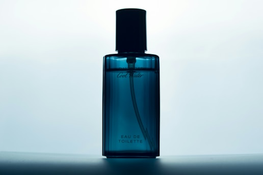 A blue glass bottle of Eau de Toilette stands against a softly lit background. The bottle has a sleek design with a black cap and subtle branding on the front.