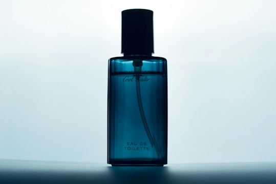 A blue glass bottle of Eau de Toilette stands against a softly lit background. The bottle has a sleek design with a black cap and subtle branding on the front.