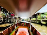 A long-tail boat with a wooden interior navigates through a canal lined with houses on stilts. Green plants, red flowers, and containers are visible on the decks of the houses. The canal water is a murky brown, and there are traditional Thai elements such as spirit houses visible.