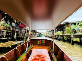 A long-tail boat with a wooden interior navigates through a canal lined with houses on stilts. Green plants, red flowers, and containers are visible on the decks of the houses. The canal water is a murky brown, and there are traditional Thai elements such as spirit houses visible.