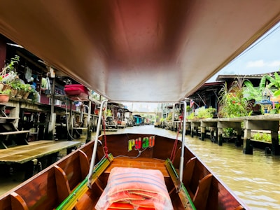 A long-tail boat with a wooden interior navigates through a canal lined with houses on stilts. Green plants, red flowers, and containers are visible on the decks of the houses. The canal water is a murky brown, and there are traditional Thai elements such as spirit houses visible.