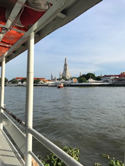 View from a boat on a river with a prominent temple spire in the background across the water. Lush greenery and traditional architecture line the riverbank, accompanied by a bright blue sky.