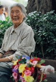 An elderly woman with a joyful expression sits outdoors surrounded by lush greenery. She is wearing a patterned shirt and looks content. In front of her is a colorful bouquet of flowers with vibrant reds, pinks, yellows, and purples.