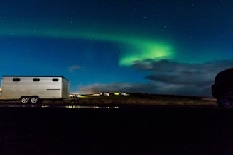 A trailer is parked on a dark roadside under a vivid display of the northern lights, which arc across a starry night sky. The scene is set on a wide-open landscape with distant lights from buildings illuminating the horizon. Clouds add texture to the night sky, complementing the green and blue hues of the aurora borealis.