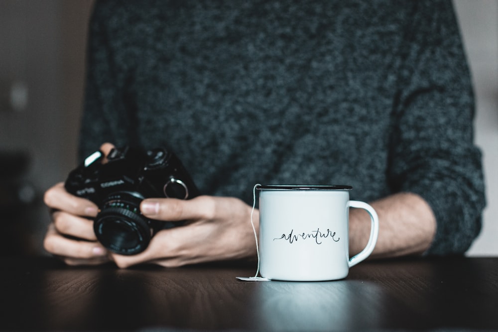 Persona che tiene la macchina fotografica vicino alla tazza di ceramica bianca sul tavolo