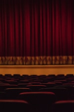 Rows of theater seats face a stage with closed heavy red curtains hanging down. The lighting is dim, creating a warm and intimate atmosphere.