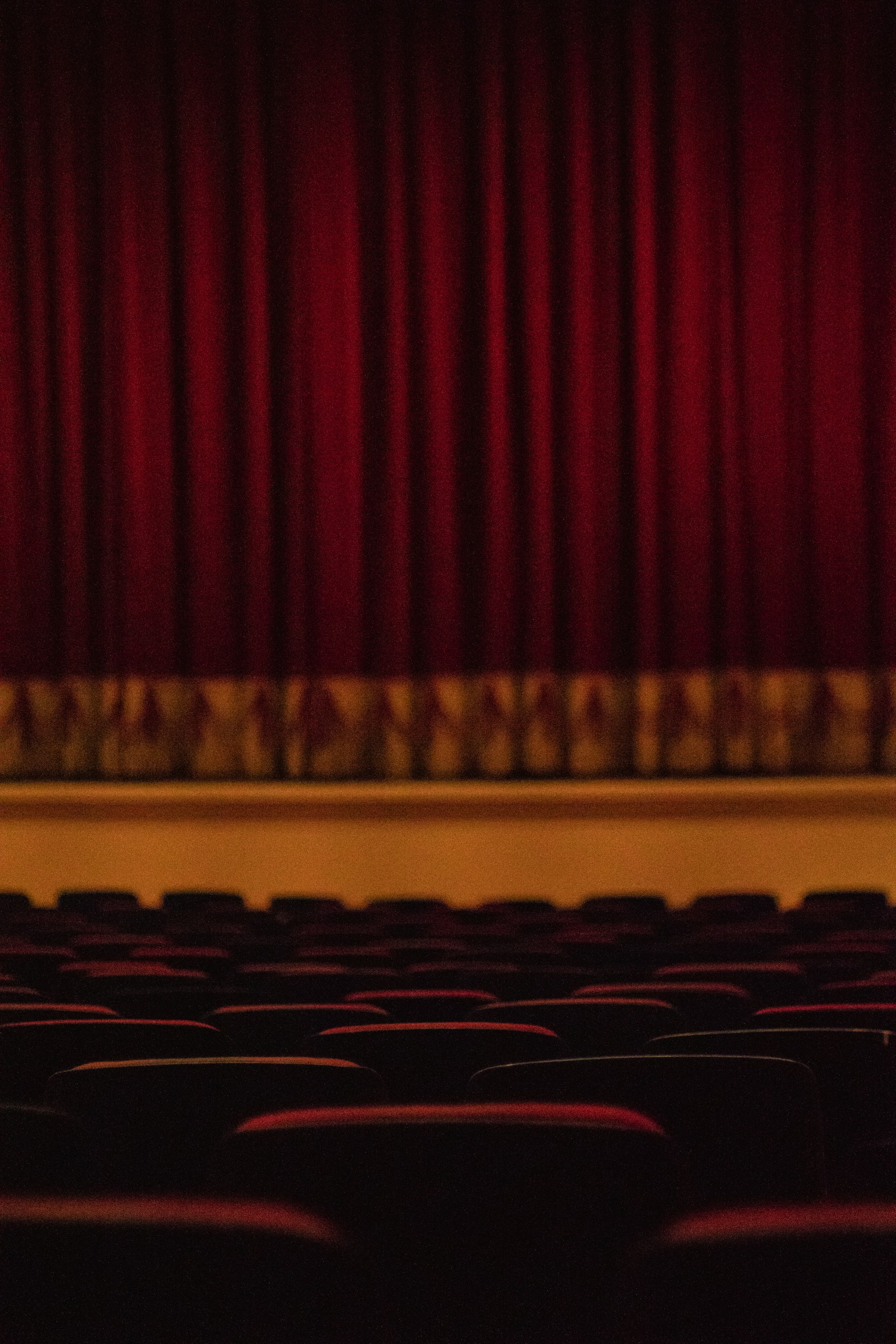 Rows of theater seats face a stage with closed heavy red curtains hanging down. The lighting is dim, creating a warm and intimate atmosphere.