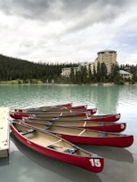 A row of red canoes is neatly lined up on a calm, turquoise lake next to a wooden dock. In the background, there is a large hotel building surrounded by a dense forest of tall, dark green pine trees under an overcast sky.