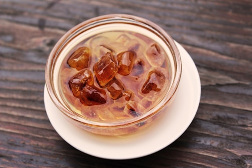 A glass bowl containing several amber-colored crystal-like sugar pieces is placed on a white saucer. The background is a rustic wooden surface with visible grain patterns.