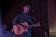 A person wearing a cap and glasses holds an acoustic guitar and sings into a microphone. The setting appears to be a stage with dim lighting, creating an intimate atmosphere. Another person is visible in the background, sitting quietly.