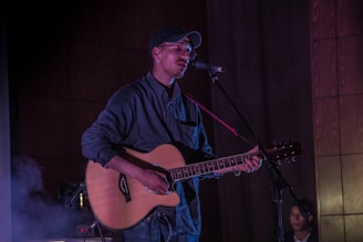 A person wearing a cap and glasses holds an acoustic guitar and sings into a microphone. The setting appears to be a stage with dim lighting, creating an intimate atmosphere. Another person is visible in the background, sitting quietly.