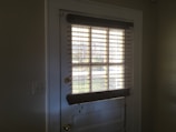 A doorway with a white door featuring glass panels covered by horizontal blinds. Sunlight streams through the blinds, casting shadows on the door and walls. The doorknob is brass-colored and a pair of light switches is visible on the left wall.