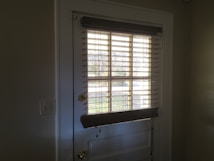A doorway with a white door featuring glass panels covered by horizontal blinds. Sunlight streams through the blinds, casting shadows on the door and walls. The doorknob is brass-colored and a pair of light switches is visible on the left wall.