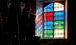 A stained glass window composed of panels in blue, red, green, and clear colors allows light to cast vibrant patterns onto an interior wall. Intricate wrought iron designs are embedded within each colored panel, creating detailed shadows across the room. Ornate metallic decorations hang nearby, contributing to the vintage ambiance.
