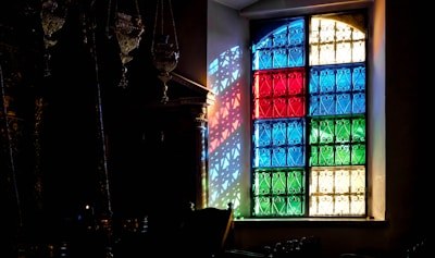 A stained glass window composed of panels in blue, red, green, and clear colors allows light to cast vibrant patterns onto an interior wall. Intricate wrought iron designs are embedded within each colored panel, creating detailed shadows across the room. Ornate metallic decorations hang nearby, contributing to the vintage ambiance.