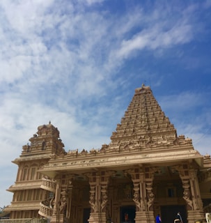 An intricately carved temple with a pyramid-shaped tower reaching towards a clear blue sky. The architecture features elaborate details, including sculpted pillars and decorative elements. The foreground shows people in traditional attire walking near the entrance.