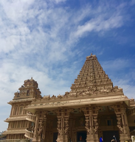 An intricately carved temple with a pyramid-shaped tower reaching towards a clear blue sky. The architecture features elaborate details, including sculpted pillars and decorative elements. The foreground shows people in traditional attire walking near the entrance.