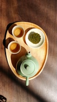 A wooden tray holds a green ceramic teapot and two small matching cups. Beside the cups, a plate contains a green decorative pastry, resembling a mooncake. Sunlight casts shadows, accentuating the earthy tones and textures of the tray and the table beneath.