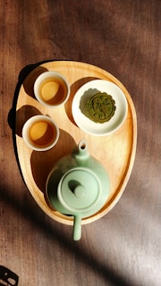 A wooden tray holds a green ceramic teapot and two small matching cups. Beside the cups, a plate contains a green decorative pastry, resembling a mooncake. Sunlight casts shadows, accentuating the earthy tones and textures of the tray and the table beneath.