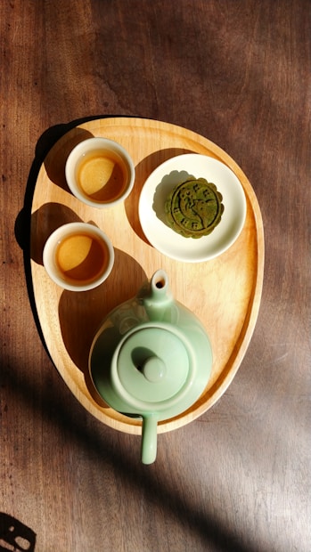 A wooden tray holds a green ceramic teapot and two small matching cups. Beside the cups, a plate contains a green decorative pastry, resembling a mooncake. Sunlight casts shadows, accentuating the earthy tones and textures of the tray and the table beneath.