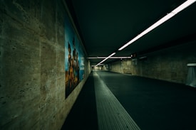 A dimly lit underground passageway with tiled walls and a large advertisement poster featuring people on the left side. The ceiling is lined with fluorescent lights that illuminate the corridor leading into the distance, and the flooring appears to be a combination of textured and smooth surfaces.