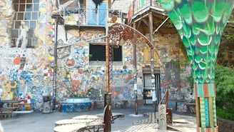 A vibrant, eclectic courtyard filled with colorful mosaic art. The walls are adorned with intricate patterns, sculptures, and various artistic elements including tiles and murals. A large, decorative metal structure and a green column with artistic designs are also present.