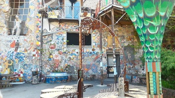 A vibrant, eclectic courtyard filled with colorful mosaic art. The walls are adorned with intricate patterns, sculptures, and various artistic elements including tiles and murals. A large, decorative metal structure and a green column with artistic designs are also present.
