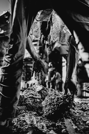 A person is captured bending over while handling rough, textured material with their hands, which are visibly soiled. The setting appears to be industrial or rugged, with blurred figures in the background suggesting activity. The image is in black and white, enhancing the gritty atmosphere.