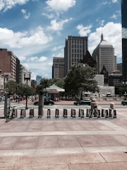 A cityscape featuring tall buildings under a partly cloudy sky. In the foreground, a row of bike-sharing stations is visible, with one bike secured. People can be seen walking in the distance, and vehicles are parked alongside the street. Trees provide green accents amidst the urban setting.