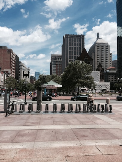 A cityscape featuring tall buildings under a partly cloudy sky. In the foreground, a row of bike-sharing stations is visible, with one bike secured. People can be seen walking in the distance, and vehicles are parked alongside the street. Trees provide green accents amidst the urban setting.